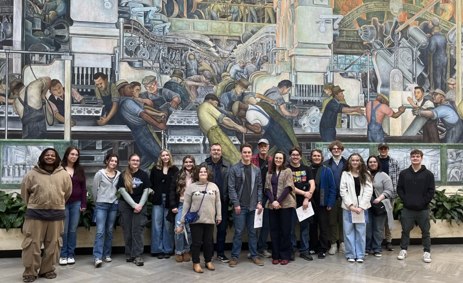 Whitchurch (left) poses at Valley’s Dec. 2025 commencement program KVCC students stand before a mural by Diego Rivera.