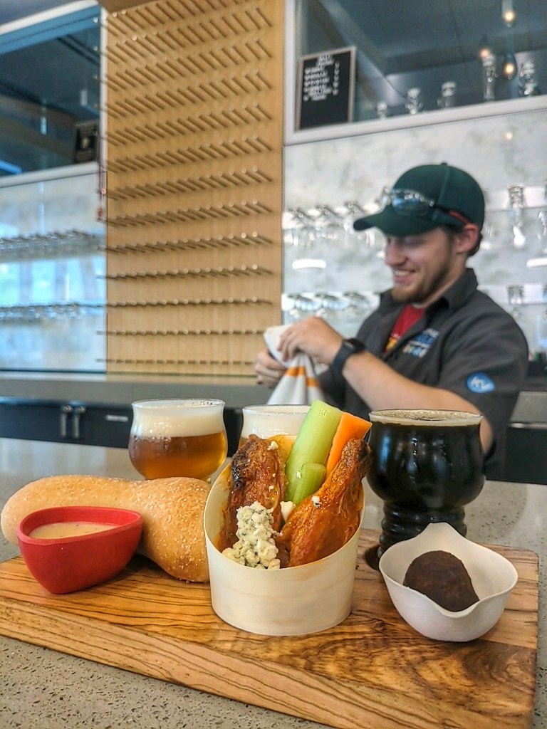 A man smiles as he dries dishes behind a counter. In the background is a great deal of glassware; in the foreground, the counter displays an assortment of food and drink.