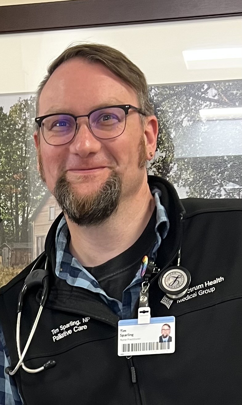 A smiling man wearing glasses and a trimmed goatee, dressed in a black vest with the text 'Tim Sparling, NP Palliative Care' embroidered on it. He also has a blue and black checkered shirt underneath and a stethoscope around his neck. A badge clipped to his vest displays his name, 'Tim Sparling,' with the title 'Nurse Practitioner.' A round medical badge and a rainbow pin are also visible on his vest. The background includes a framed picture of a rural house and trees.