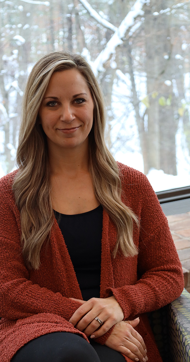 A woman with long, wavy blonde hair sits in a relaxed pose, wearing a rust-colored knit cardigan over a black top. She has a subtle smile and her hands are clasped in her lap, with a wedding ring visible. Behind her is a window showcasing a snowy outdoor scene with trees and branches.