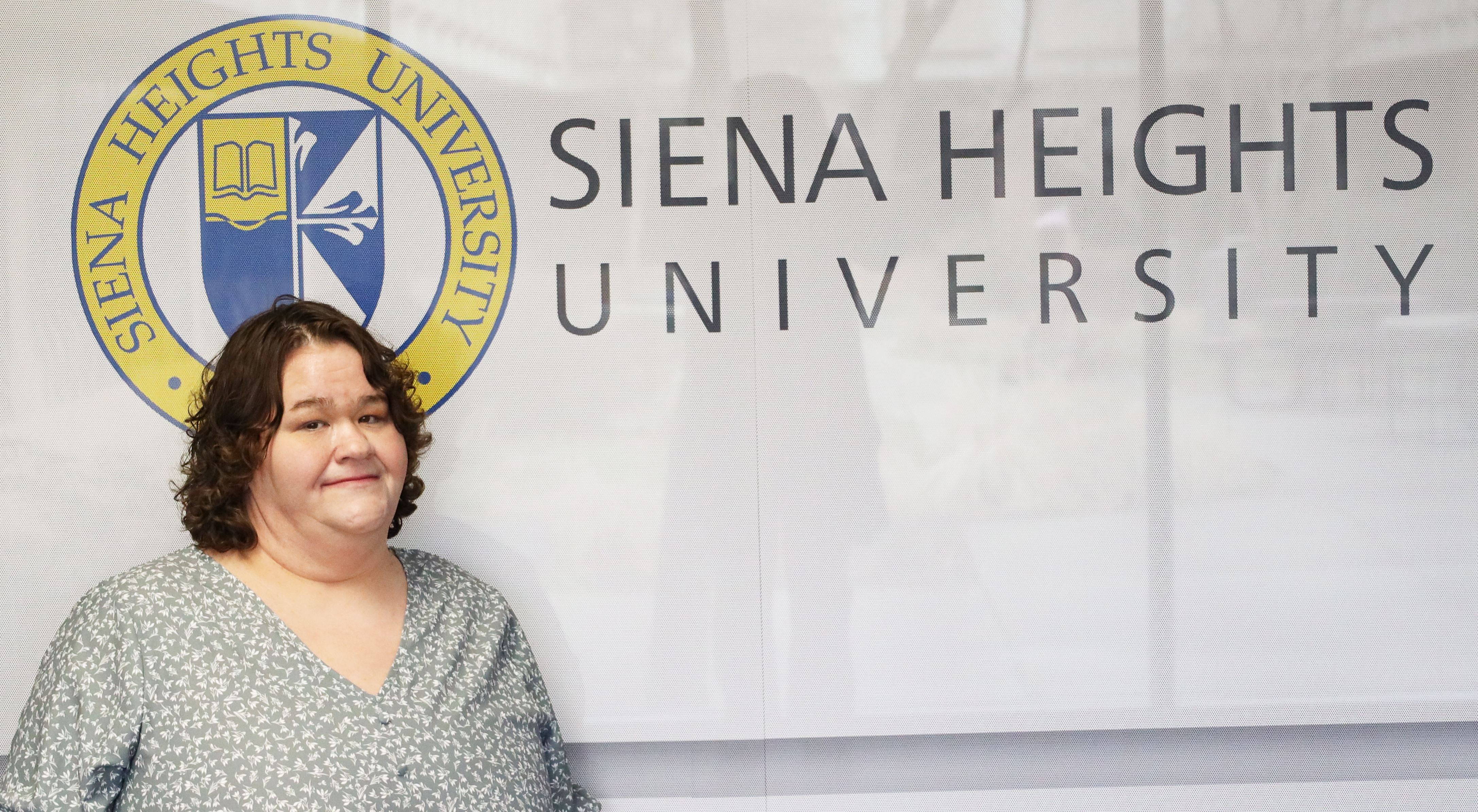 A woman with short dark hair and a smile, wearing a floral print top, stands before a sign displaying the blue and gold logo for Siena Heights University.