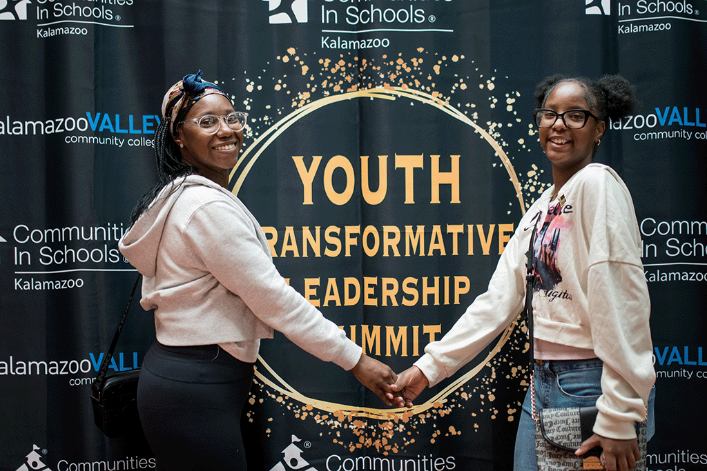 Two smiling young women stand in front of a black and gold event backdrop that reads "Youth Transformative Leadership Summit." The backdrop also features the logos for Kalamazoo Valley Community College and Communities in Schools of Kalamazoo. The women are holding hands and facing the camera, symbolizing unity and partnership.